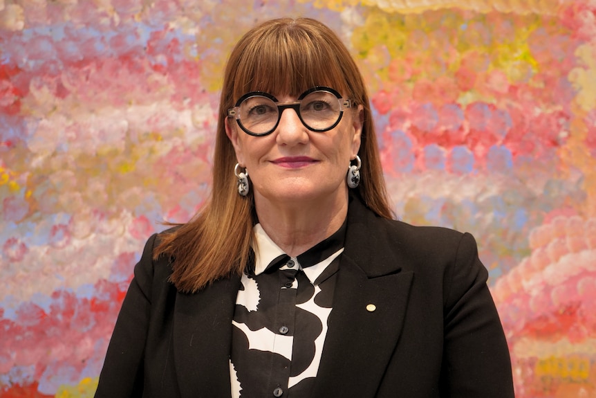 A woman with rounded glasses standing in front of a bright painting looking into the camera.