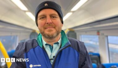 Paul is wearing a Northern uniform, standing in a railway carriage, smiling at the camera.