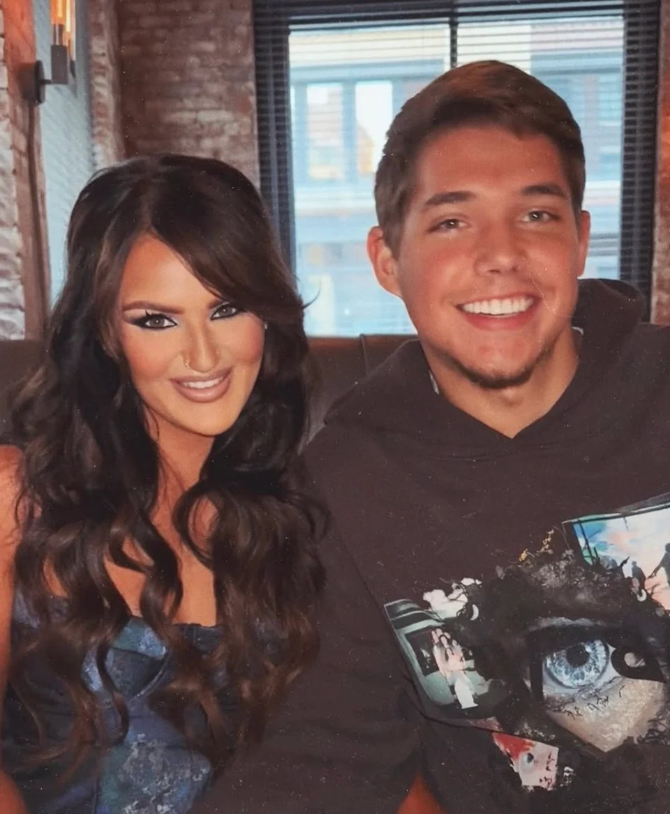 Two people smiling indoors; the person on the left has long hair and is wearing a strapless outfit; the person on the right is in a casual sweater