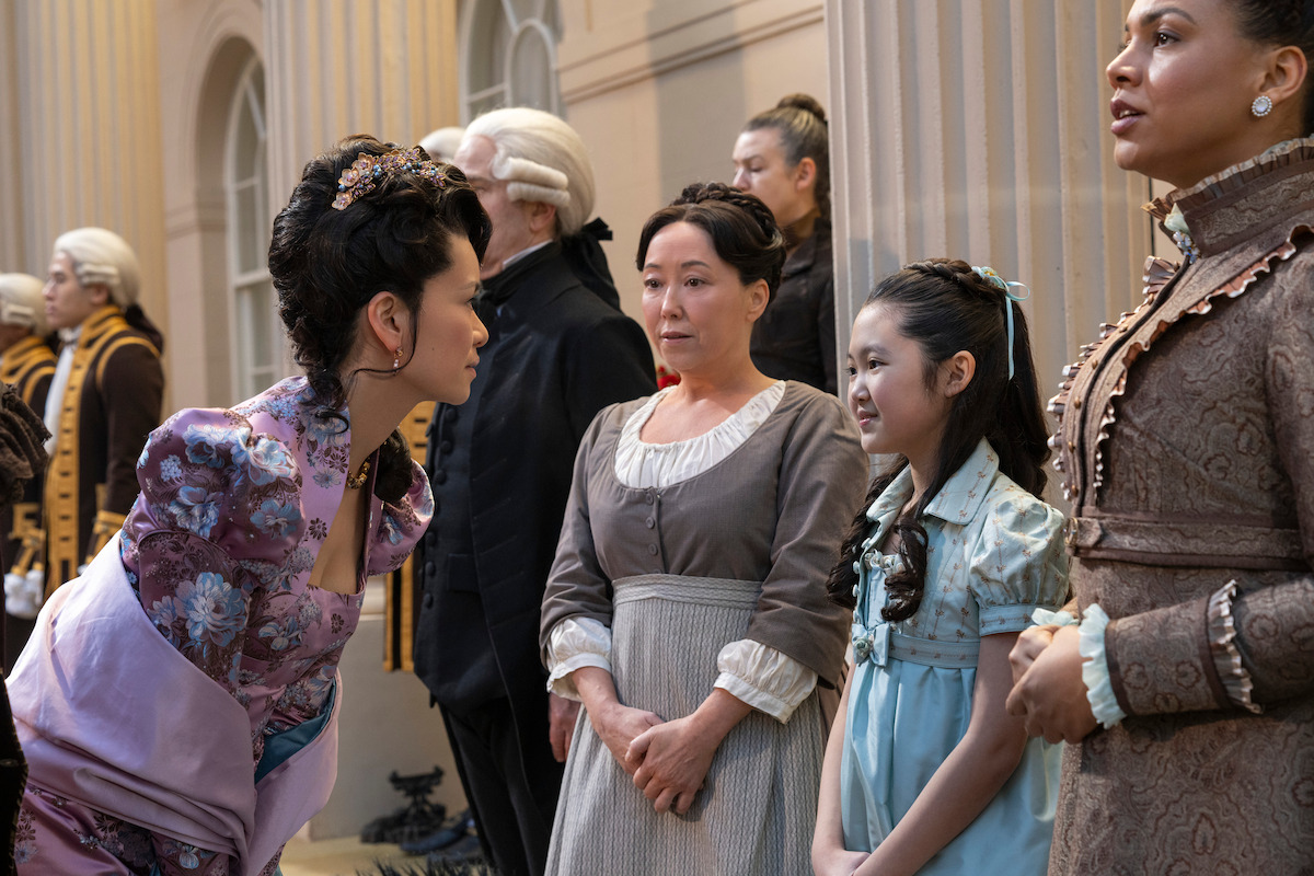 Women and a girl in elaborate historical costumes stand in a grand, ornate hall with tall columns and arches, surrounded by others in period clothing, suggesting a formal gathering or event.