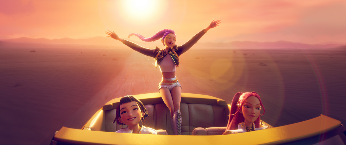 Three animated young women ride in a yellow convertible across a desert at sunset; one stands up joyfully with arms raised while the other two smile in the front seats, surrounded by warm, glowing light.