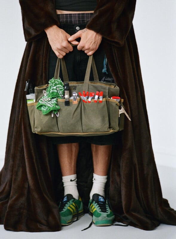 A person wearing a dark brown robe, green Adidas sneakers, and white Nike socks holds an olive green tote bag filled with red markers, a green scarf, a lock, and other items. Their face is not visible.