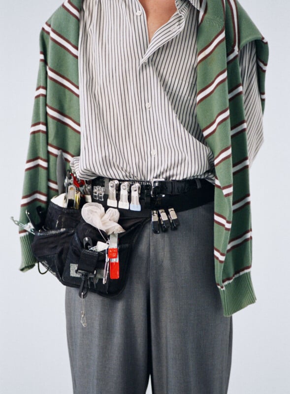A person wearing a green and white striped sweater over a striped shirt and gray pants has a utility belt with various clips, tools, and accessories attached to it around their waist.