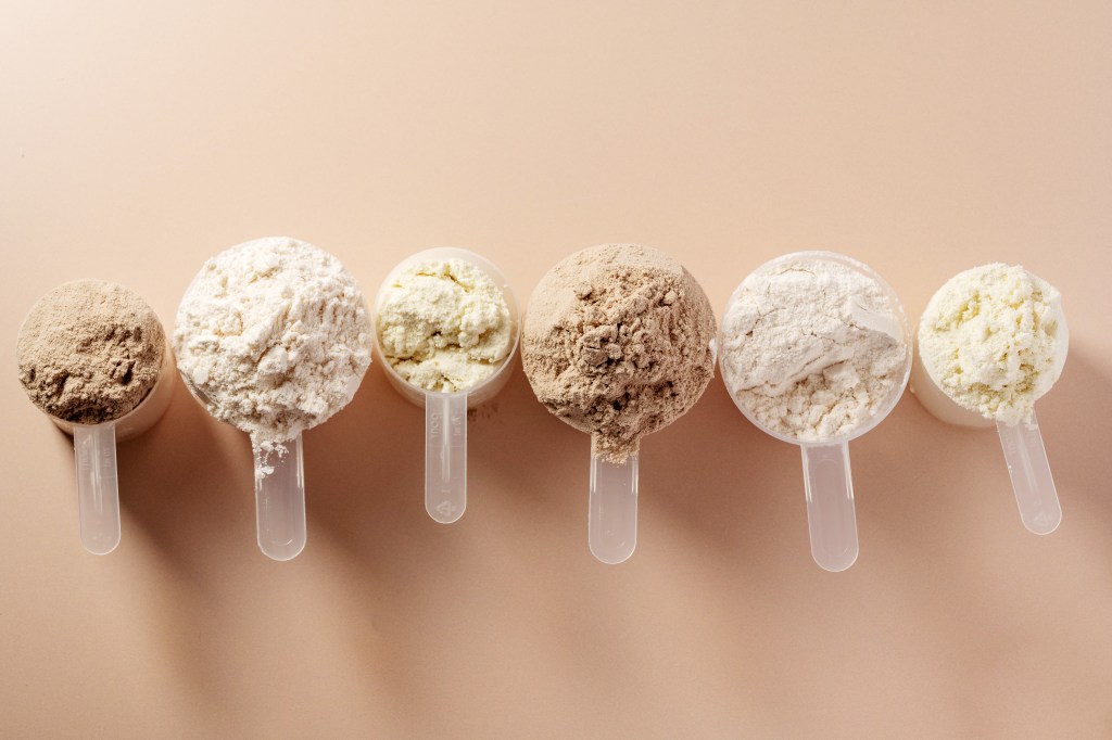 Six scoops of protein powder in various shades of brown and off-white, arranged in a row on a beige background.