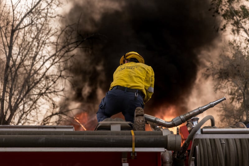 A firefighter in yellow protective gear stands atop a fire truck, aiming a hose at a large blaze with thick black smoke rising among trees.