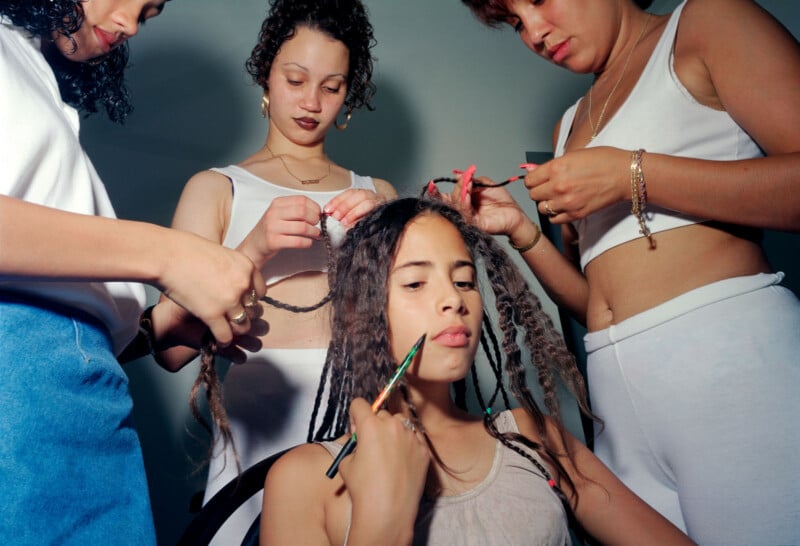 Three women work together to style another woman’s hair, braiding and trimming her curls. The seated woman looks forward while holding a comb to her chin. All four wear casual, light-colored clothing.