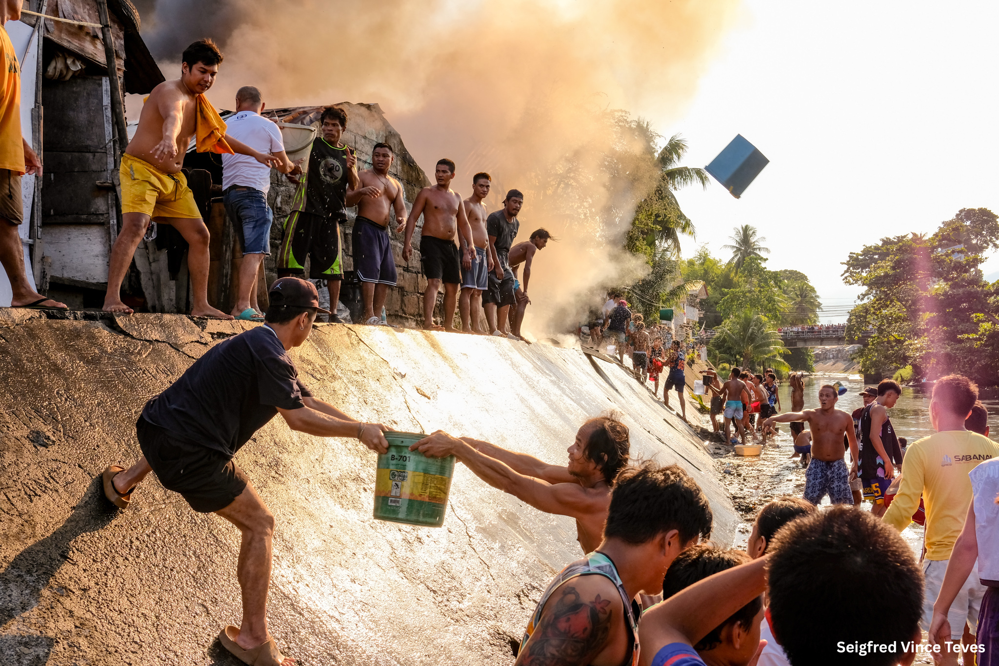 People forming a human chain pass buckets of water to fight a fire, with smoke billowing in the background. The mood is urgent and cooperative