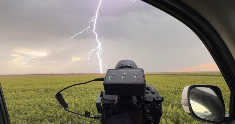 A camera set up on a tripod inside a car captures lightning striking in a field under a cloudy sky, with green crops stretching to the horizon.