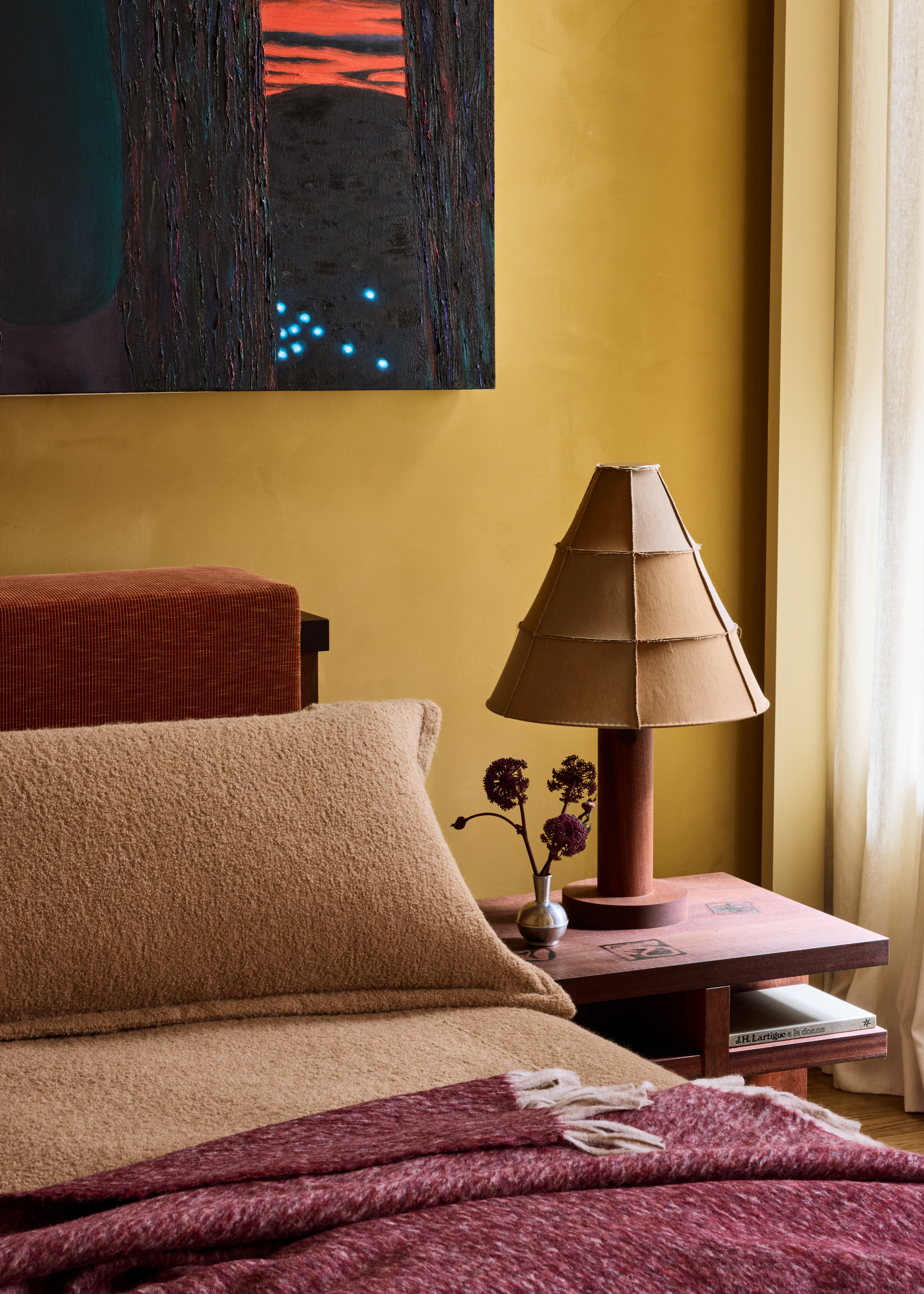 yellow bedroom with large artwork, timber side table with cool lamp, and bud flower vase, orange corduroy headboard, with wool tan pillowcase and bedding