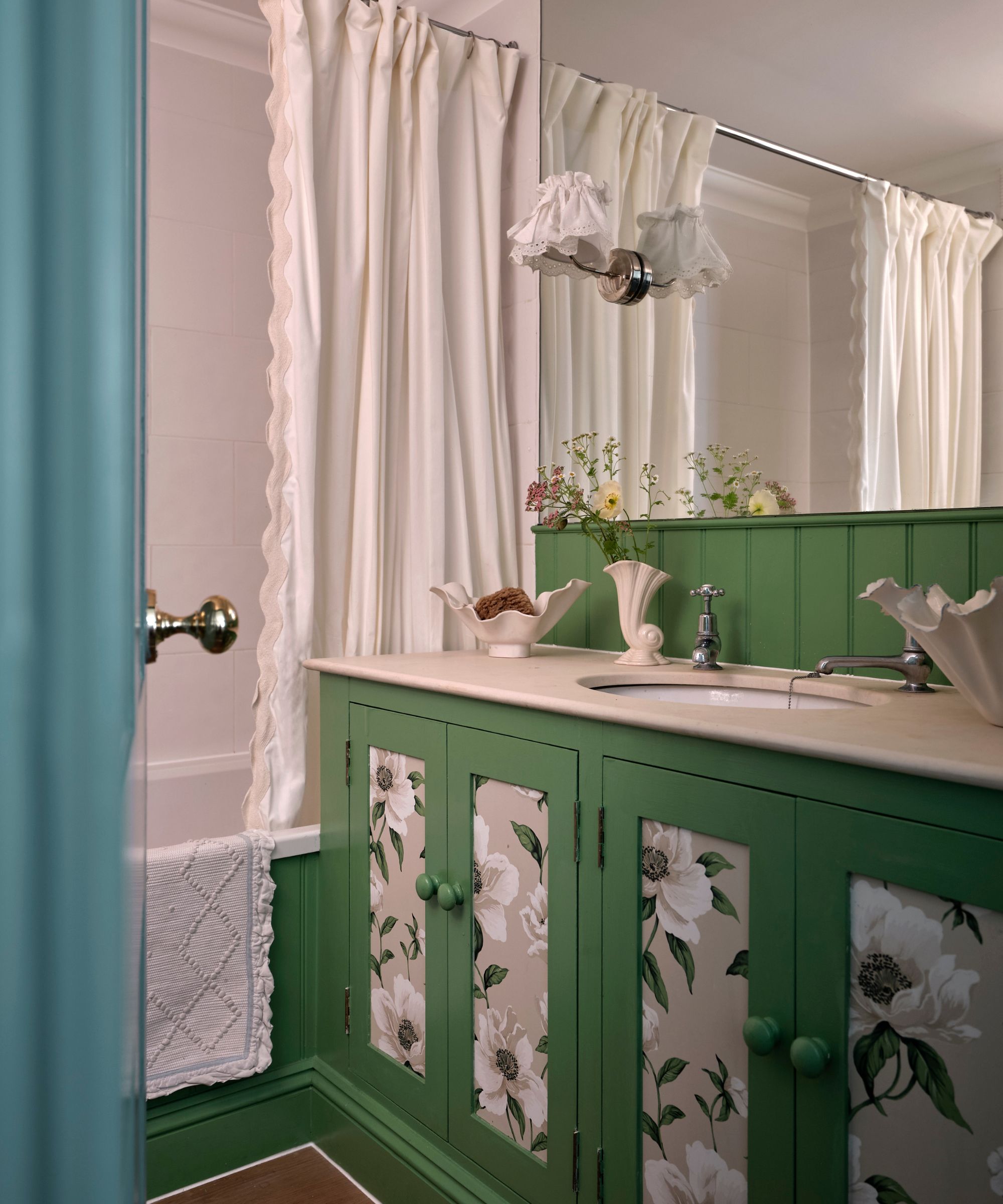A vibrant green bathroom vanity with floral-patterned door panels and a white stone countertop. A white scalloped shower curtain and matching shell-shaped dishes add a vintage touch.