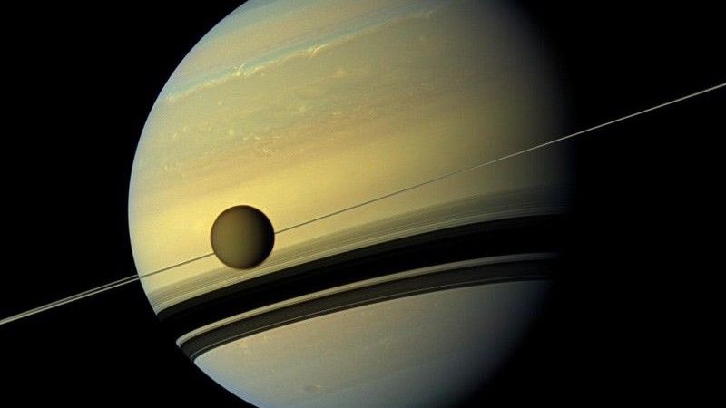 A view of Saturn with its rings creating a shadow on its body. A moon is in the foreground.