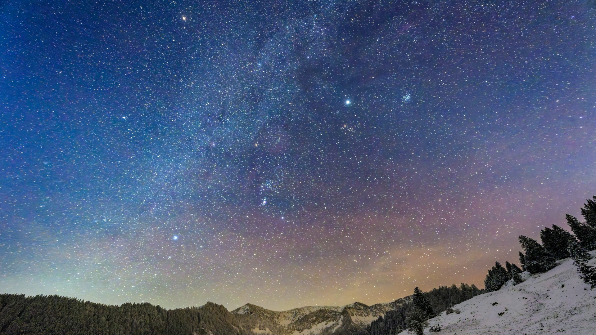 A picture of the glowing band of the Milky Way travelling diagonally from lower left to upper right through a starry night sky above a snowy, hilly landscape partially covered in evergreen trees.