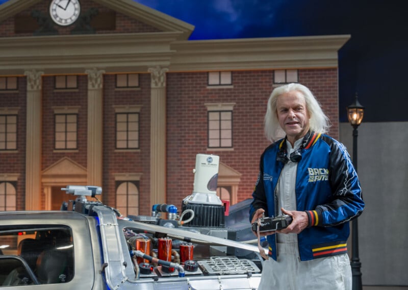 An older man with white hair wearing a blue "Back to the Future" jacket stands next to a DeLorean car, in front of a building with a large clock, resembling a scene from the film "Back to the Future.