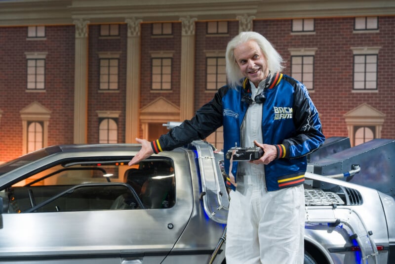 An older man with white hair, dressed in a blue "Back to the Future" jacket and white pants, stands smiling next to a silver DeLorean car, gesturing towards it with one hand. A building facade is in the background.