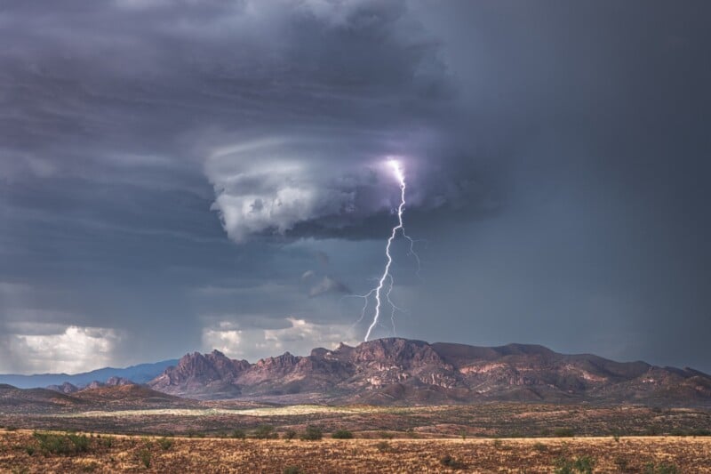 A bright lightning bolt strikes from a swirling dark cloud above rugged mountains and a dry, open plain under a stormy sky.