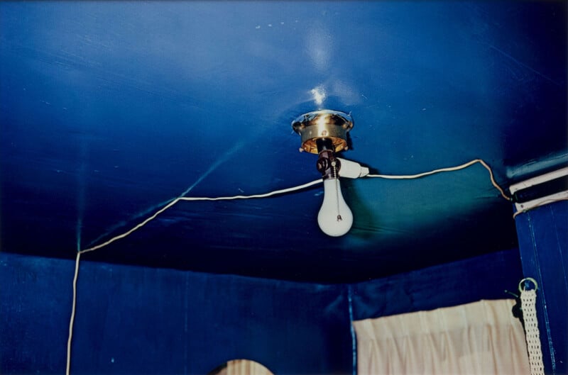 A single exposed light bulb hangs from a ceiling fixture in a corner of a room with deep blue walls and ceiling. White cords run along the ceiling, and cream-colored curtains cover windows below.