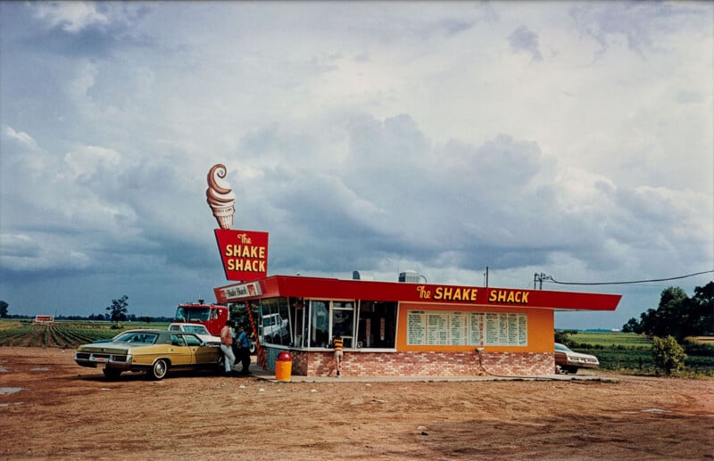 A vintage roadside Shake Shack stands under a cloudy sky, with a large ice cream cone sign on the roof. Cars are parked outside, and farmland stretches into the distance behind the small diner.