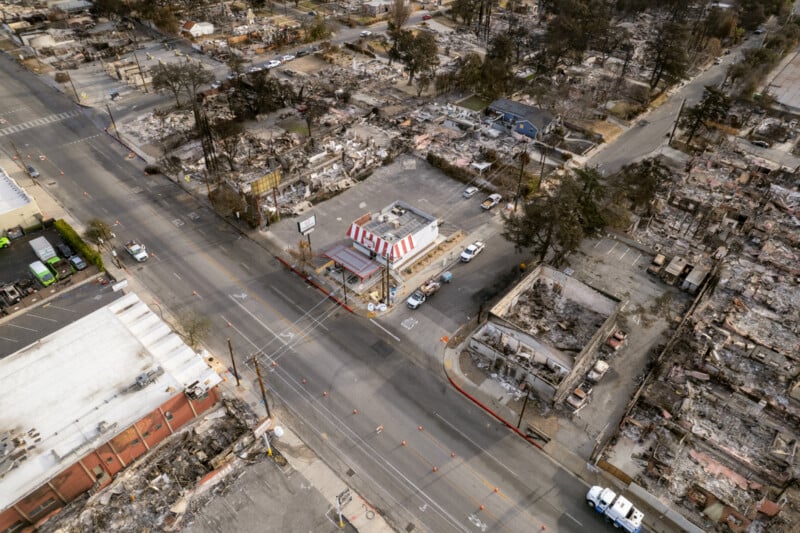 Aerial view of a fast-food restaurant with a red and white striped roof standing intact amid widespread destruction; surrounding buildings and homes are reduced to rubble from a recent fire.