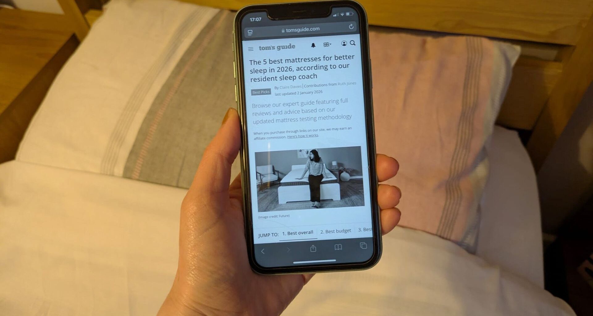 This image shows a hand holding an iPhone showing the Tom's Guide best mattress page in grayscale. In the background is a bed and pillows