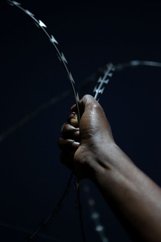 A close-up of a hand gripping a strand of razor wire against a dark background, with dramatic lighting highlighting the hand and the sharp edges of the wire.