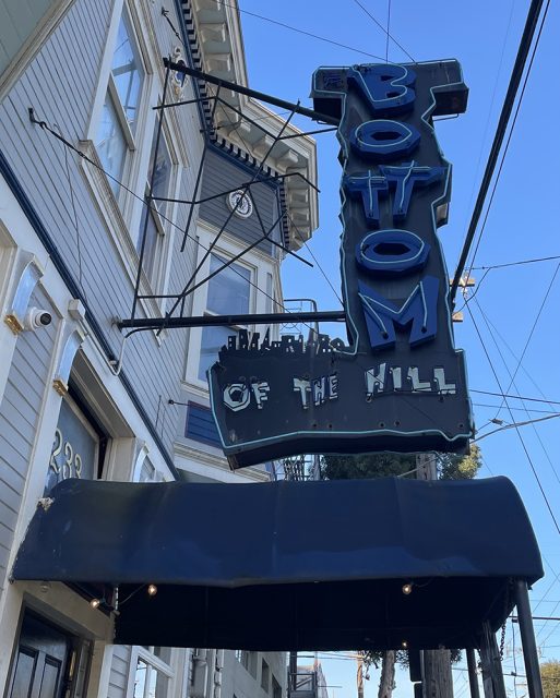 Large blue neon sign reading "Bottom of the Hill" hangs above a black awning outside a light gray building with white trim.