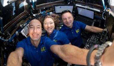 Three astronauts in blue shirts smile and pose for a selfie inside a spacecraft, surrounded by control panels, screens, and technical equipment. Bright light comes through the windows behind them.