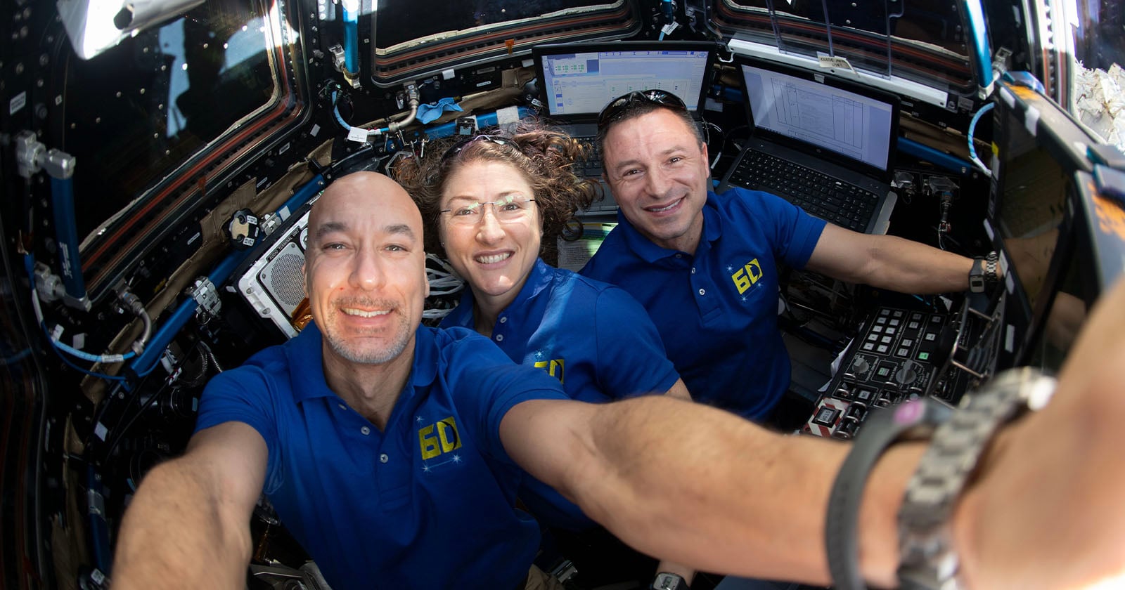 Three astronauts in blue shirts smile and pose for a selfie inside a spacecraft, surrounded by control panels, screens, and technical equipment. Bright light comes through the windows behind them.
