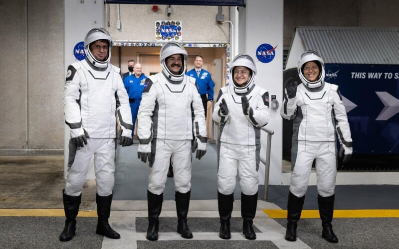Four astronauts in white SpaceX spacesuits stand side by side, smiling and posing for a photo outside a NASA building. Several people in blue uniforms stand in the background near NASA signs.
