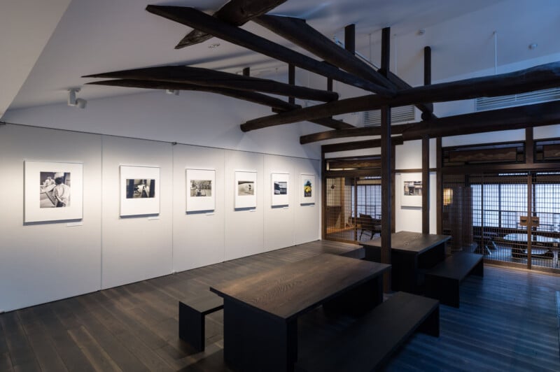 A modern gallery room with dark wooden floors and beams, displaying black-and-white photographs on a white wall. A long wooden table with benches is centered below the exposed ceiling structure. Warm lighting enhances the cozy atmosphere.