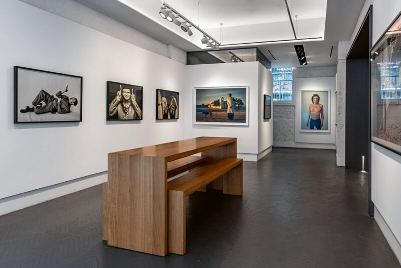 A modern art gallery with white walls displays framed photographs. A wooden table with benches sits in the center, and large windows let in natural light from the right side of the room.