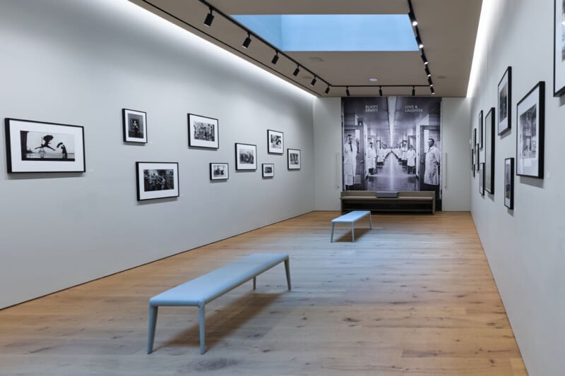 A modern art gallery with wooden floors, white walls, and black-and-white photographs displayed in black frames. Two gray benches are placed along the center, and a skylight brightens the space.