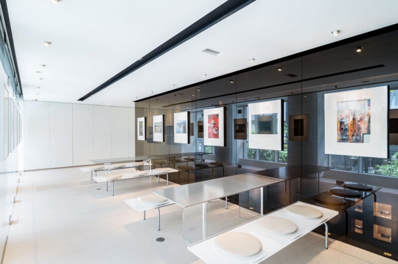 A modern, minimalistic dining area with long metal tables, white benches, and framed artwork displayed on a glossy black wall. Large windows let in natural light, brightening the clean, open space.