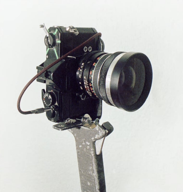 A vintage black camera with a large lens is mounted on a metal bracket against a plain light background. A cable release is attached to the camera. The metal bracket appears worn and scratched.
