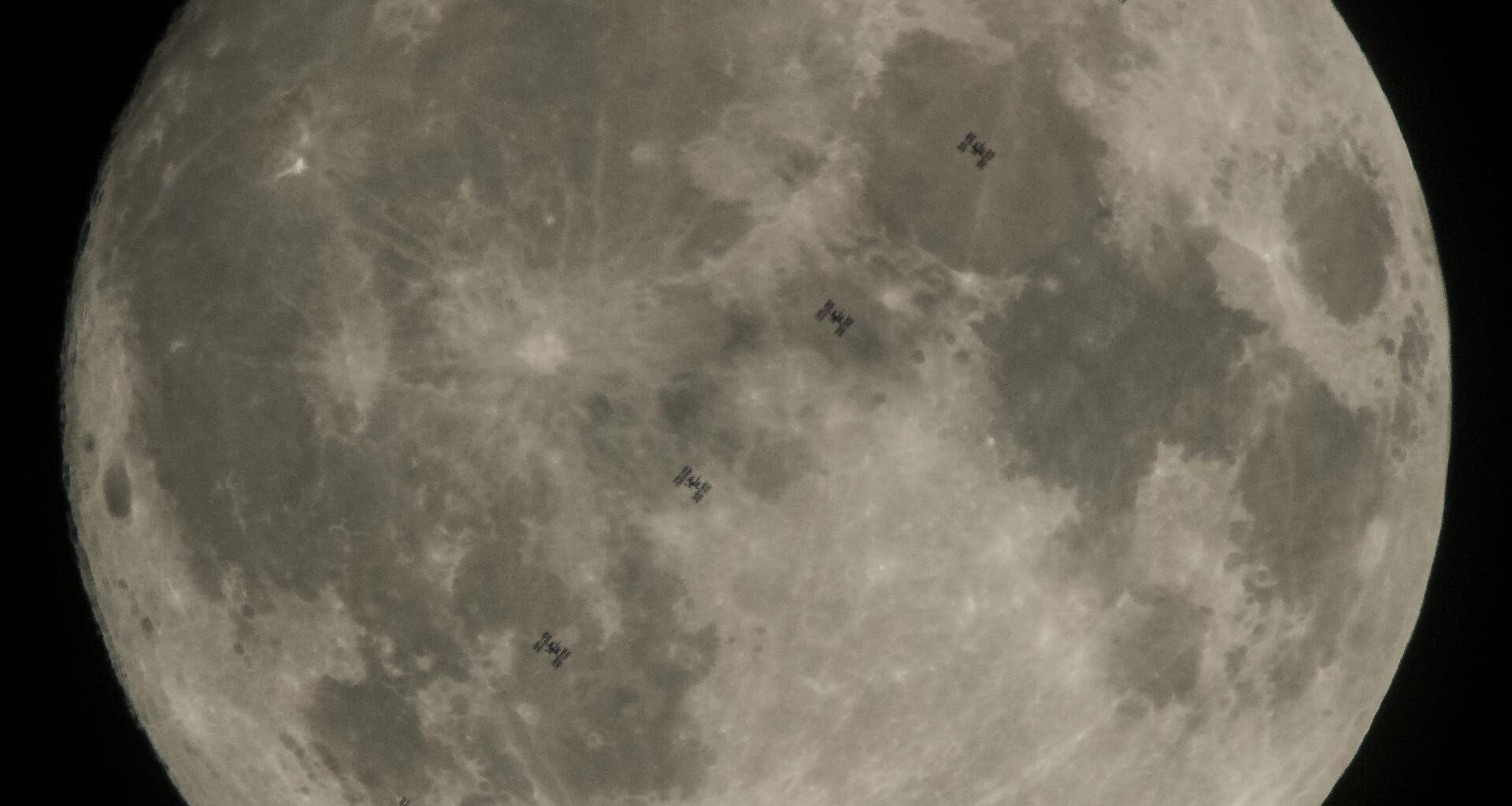 The full Moon fills the frame against a black sky, with multiple small silhouettes of the International Space Station captured as it transits across the Moon’s face.