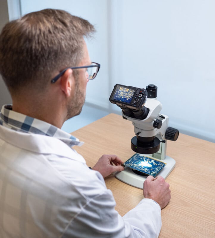 A man in a white lab coat examines a circuit board under a digital microscope on a wooden table, with a display screen showing a close-up of the board.