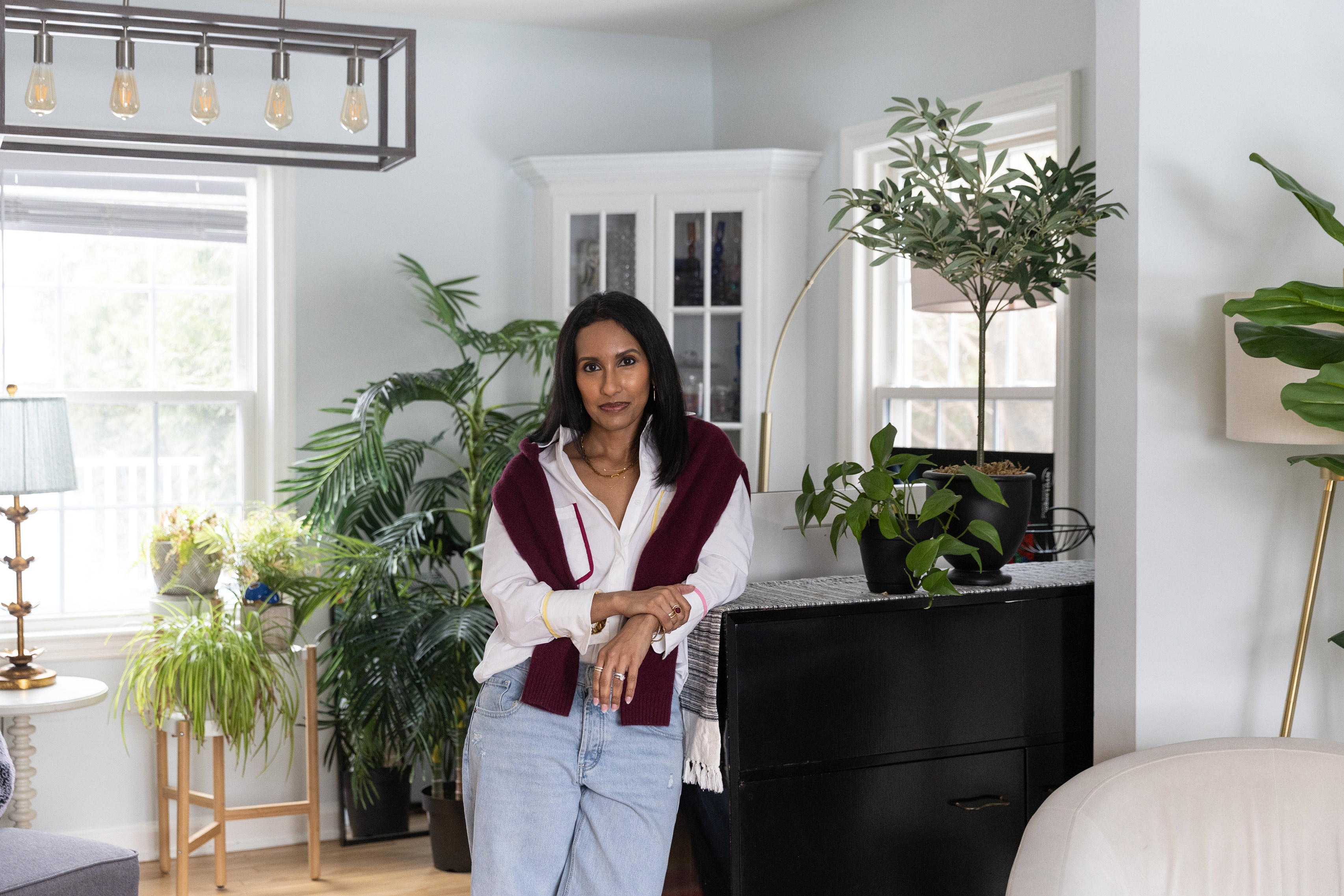 Sadia Zapp stands in her living room. Various potted plants and a china cabinet are behind her.