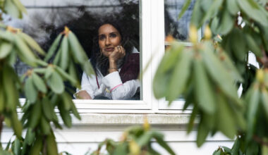 A woman looks out her window. Blurred greenery is seen in the foreground.