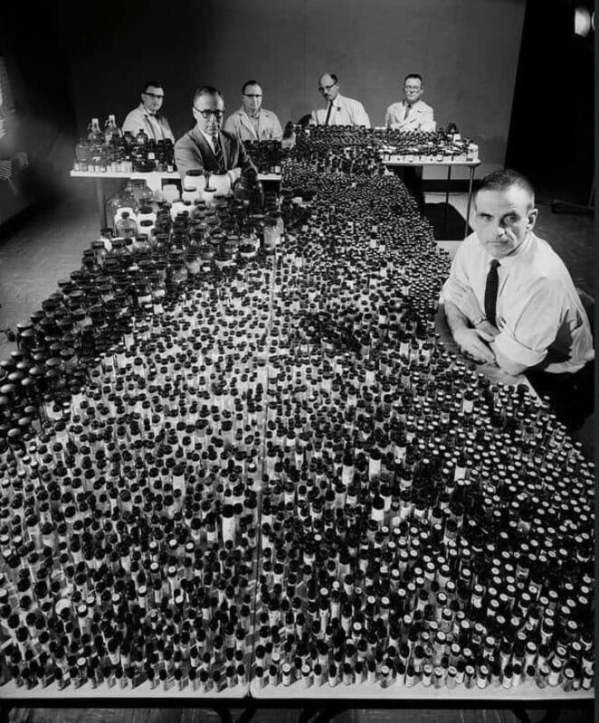 A black-and-white photo showing six men in white shirts and ties sitting behind a large table covered with hundreds of small bottles and containers arranged in neat rows.