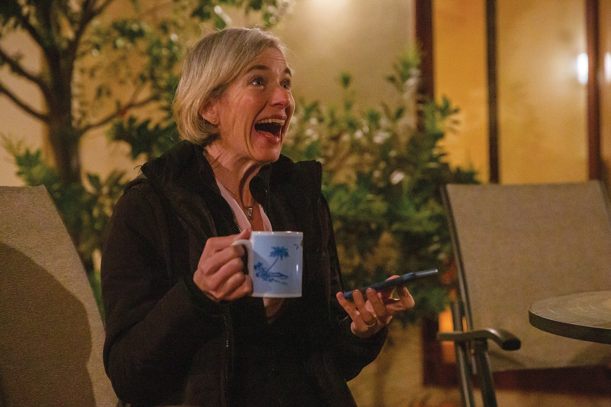 A woman wearing a coat in an outdoor setting with nighttime lighting smiles widely while holding a cell phone and a blue mug.