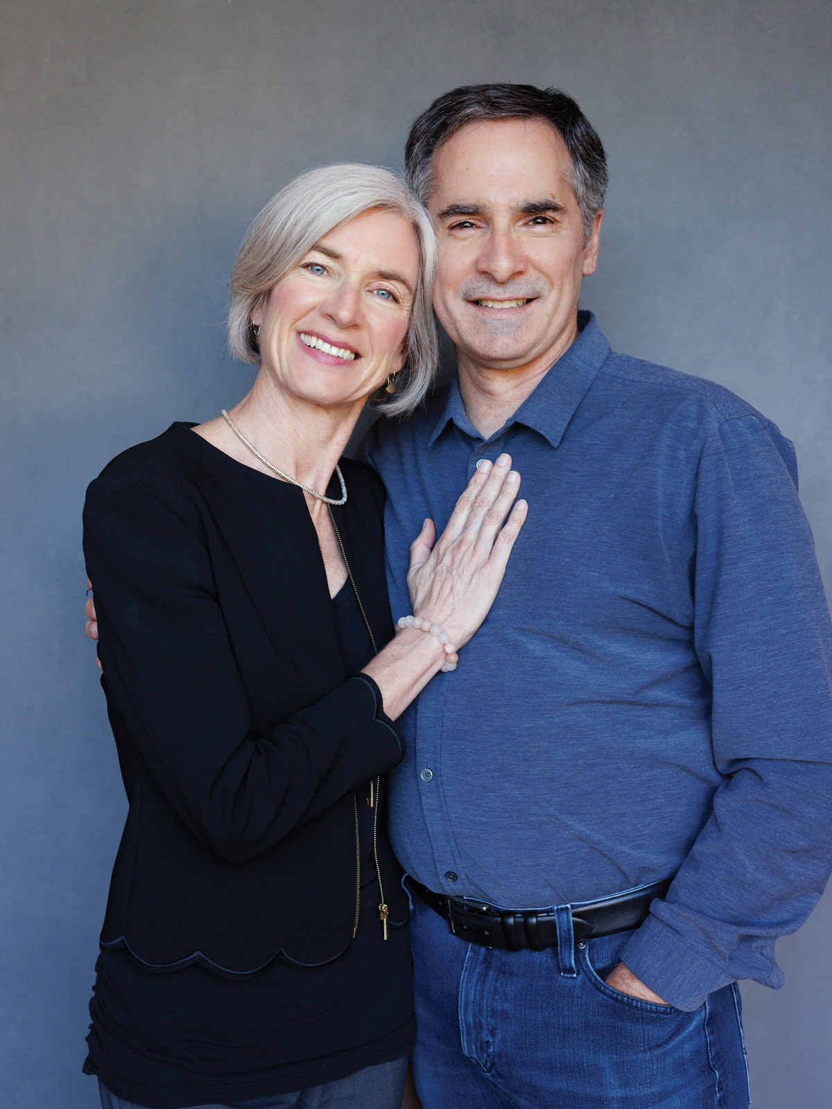 A woman in a black shirt stands with her hand on the chest of a man wearing a blue button-down shirt and jeans as both smile at the camera.