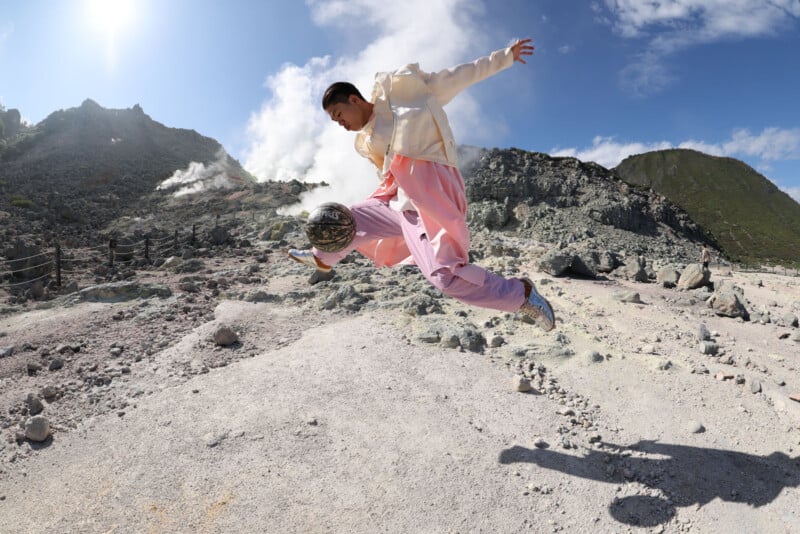 A person in pastel clothing jumps in midair while kicking a soccer ball, set against a rocky, mountainous landscape with a bright blue sky and scattered clouds.