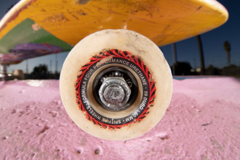 Close-up view of a skateboard wheel and truck on a pink concrete surface, with the yellow deck visible above and a blurred outdoor background.