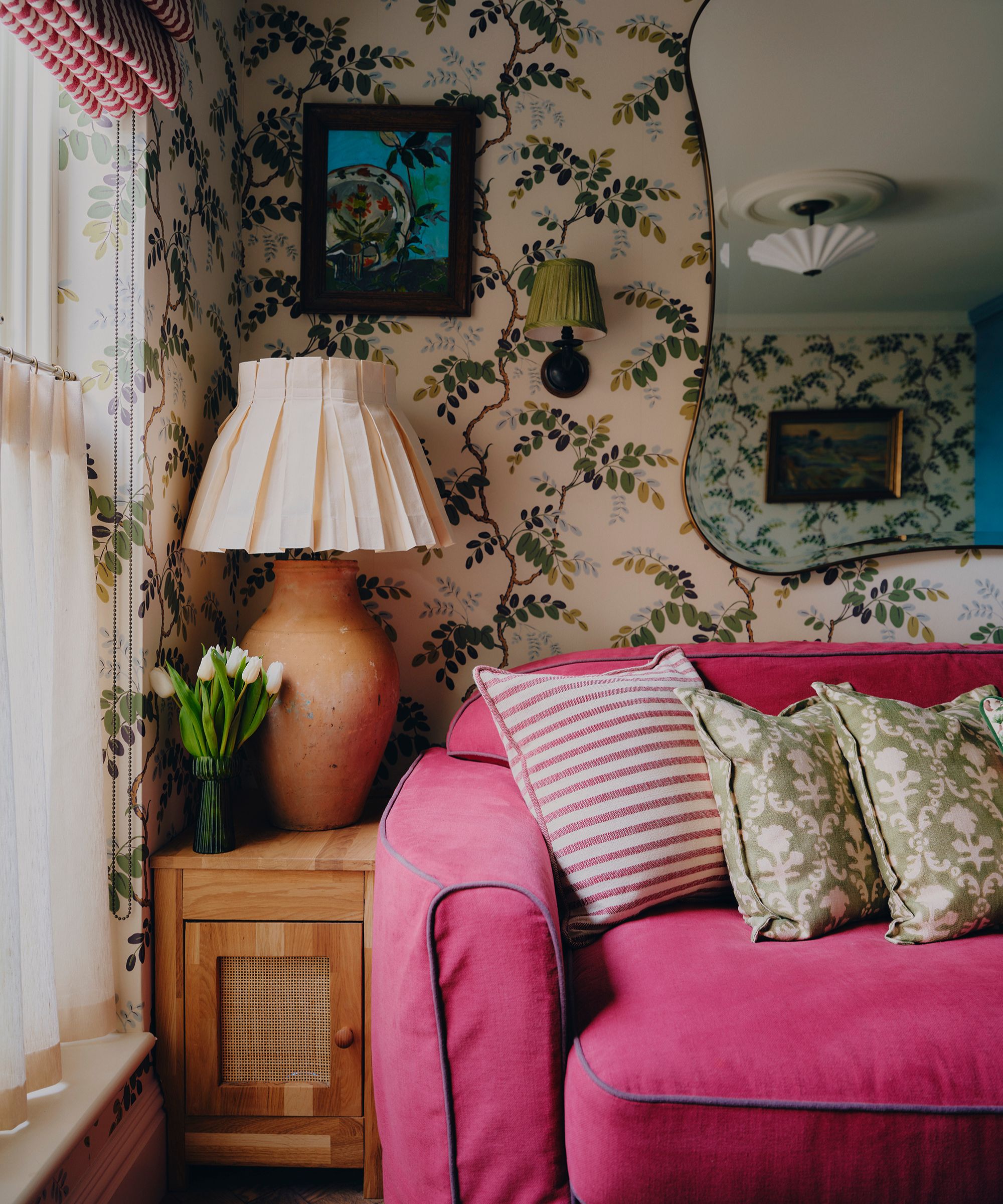 a floral wallpapered living room with a pink sofa, large italian mirror, rattan wooden cabinet, and layered art and lighting