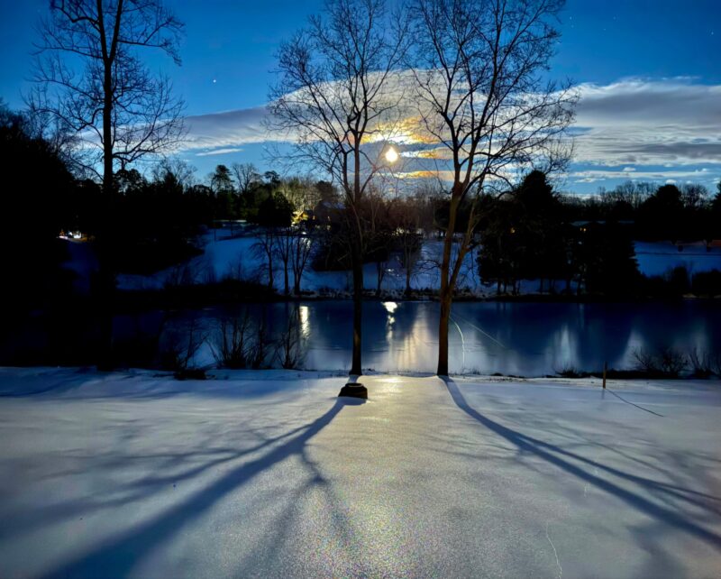 Full Snow Moon: The moon, ringed by clouds tinged with orange, among the bare branches of trees. A snowy field runs down to a creek.