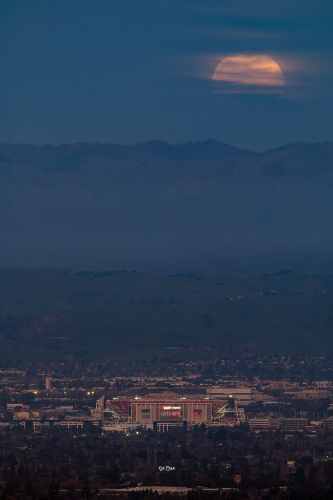 Huge orangish moon behind some clouds, over a mountainous horizon, with a city in the foreground.