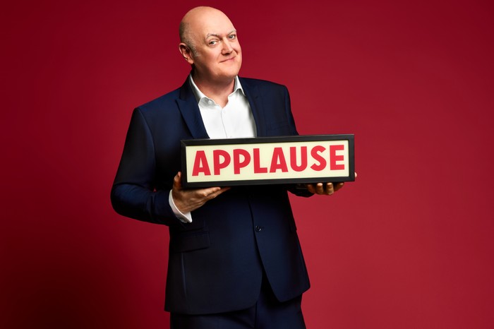 Dara Ó Briain in a navy suit smiles while holding an illuminated “APPLAUSE” sign against a solid red studio background.