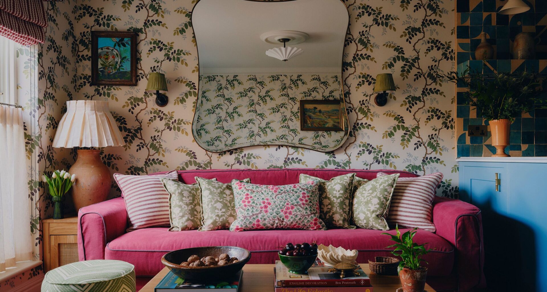 an eclectic, small living room in london with patterned wallpaper and a large dark pink couch with patterned pillows