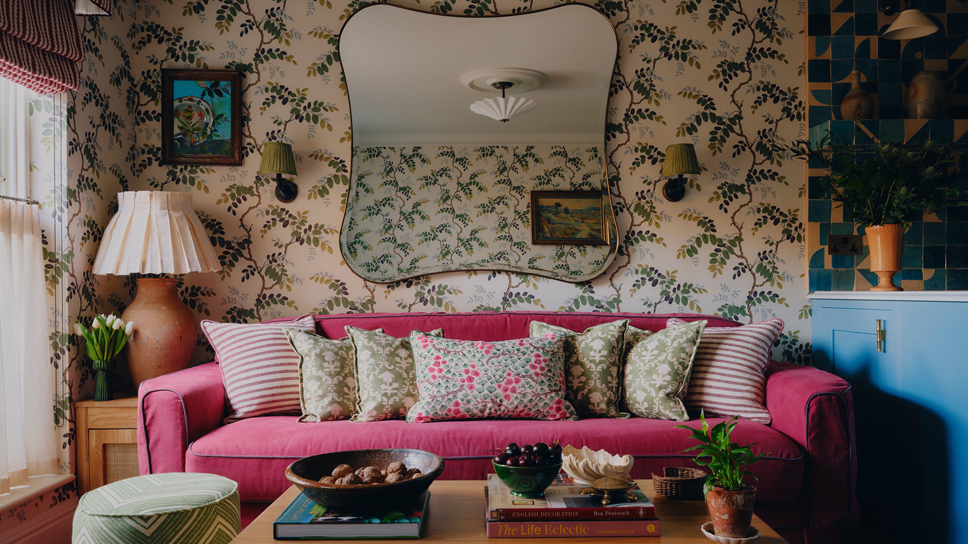 an eclectic, small living room in london with patterned wallpaper and a large dark pink couch with patterned pillows