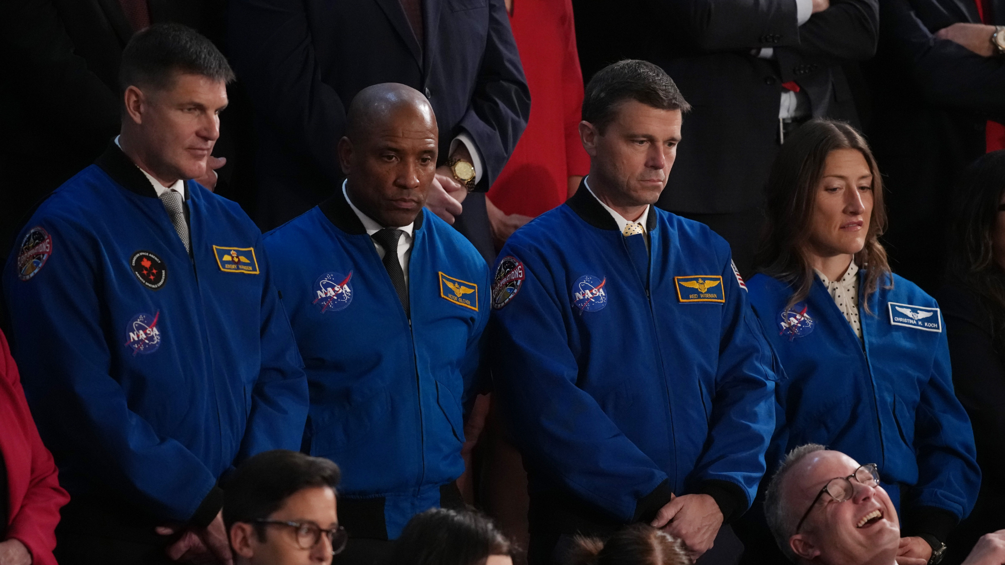 three men and one women, all wearing blue flight suits, stand solemnly in a crowd at a fancy event