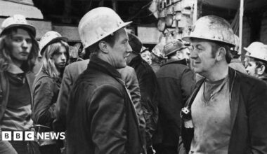 A black and white photograph of a group of men with hard hats on and in worn, dirty clothes. McGuinness stands on the right talking to another man. He has brown hair with large sideburns.